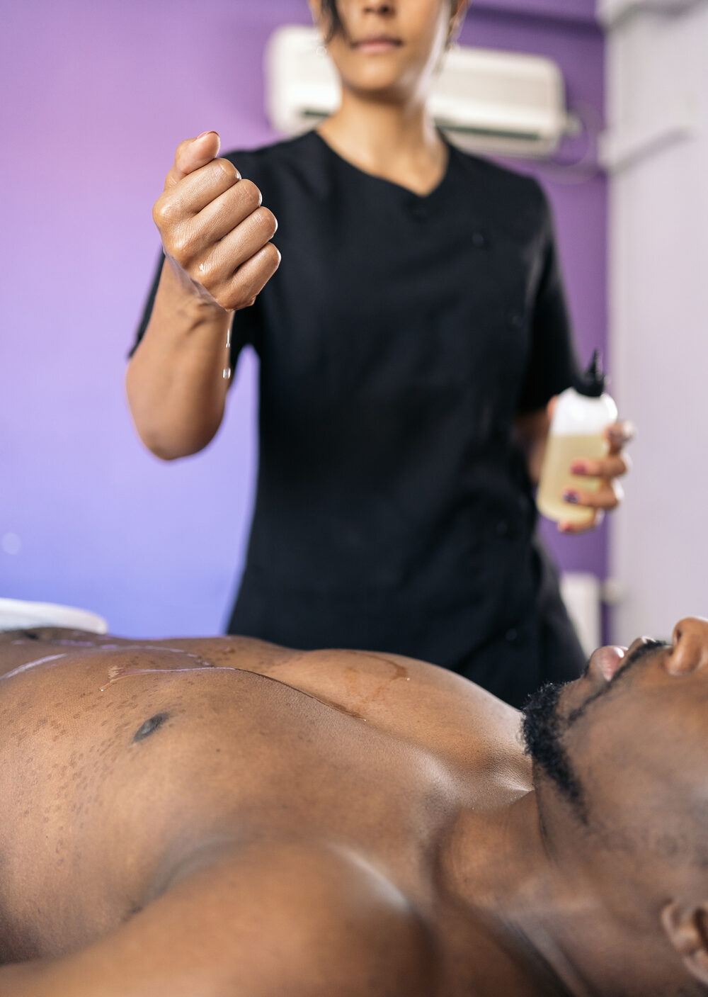 Female spa worker wearing black work clothes giving back massage to unrecognized african client and using oil.